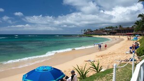 A beach with clear turquoise water, a sandy shore, and a few people. There are umbrellas and a building in the background. - Kiahuna Plantation Ocean View with AC, REMODELED, 1 Bedroom, Sleeps 4 (Poipu)