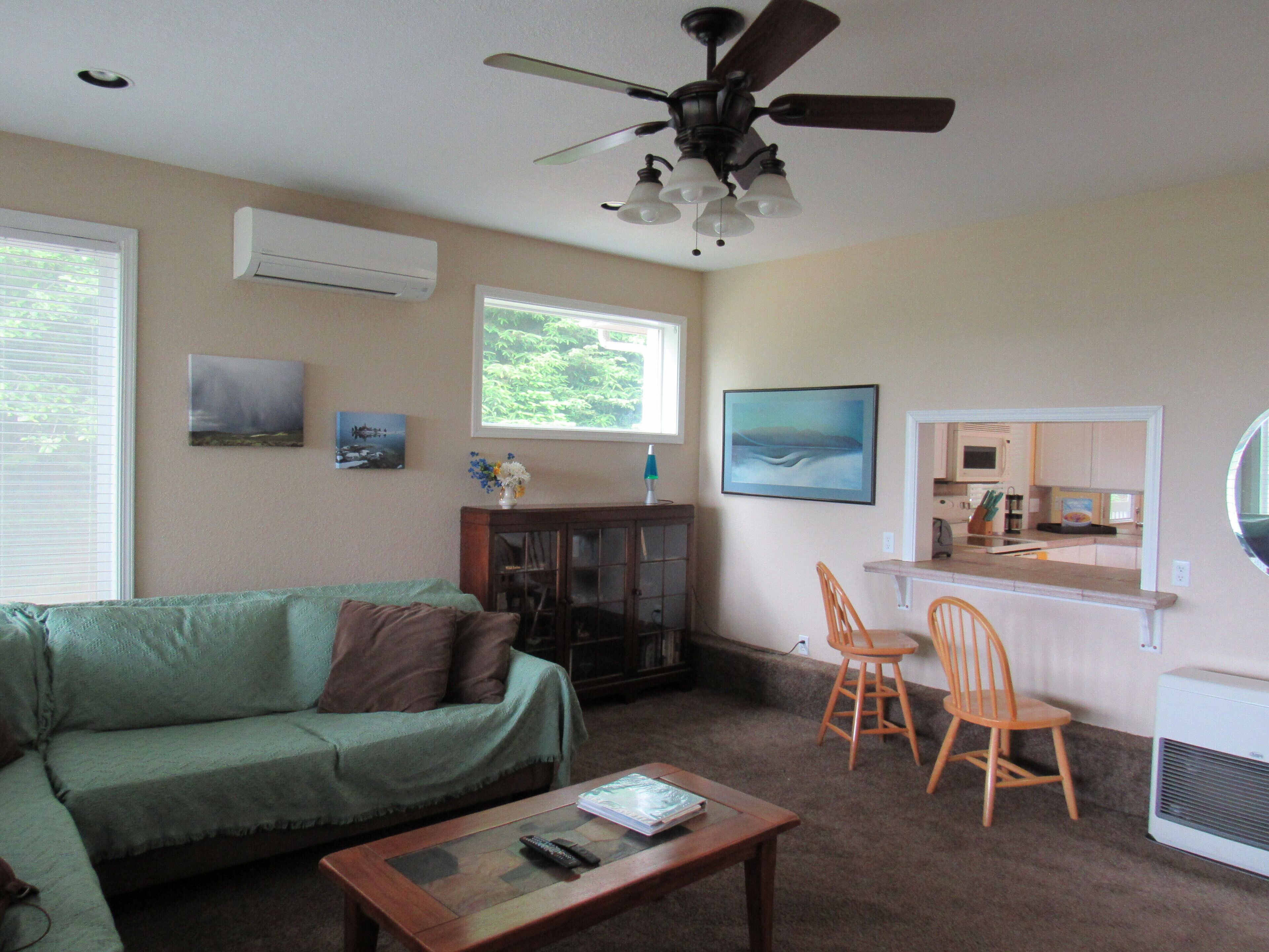 A living room with a green couch, a wooden coffee table, a ceiling fan, and a kitchen area visible through an open door.