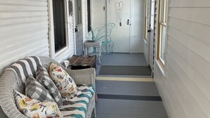 Un porche cubierto con un sofá de mimbre, una silla de metal y una puerta decorada con un adorno de corazón.