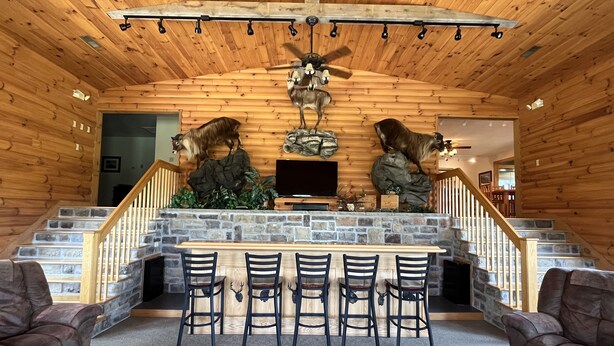 A cozy cabin interior with a stone fireplace, wooden ceiling, and a dining area with chairs.