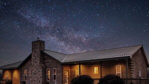 A log cabin with a stone chimney, wooden exterior, and a well-lit front porch under a starry night sky.