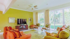 A living room with orange and green sofas, a television, and a large window with curtains. - Classic Avondale  Private  Country Club ~ Gated community front gate security (Palm Desert)