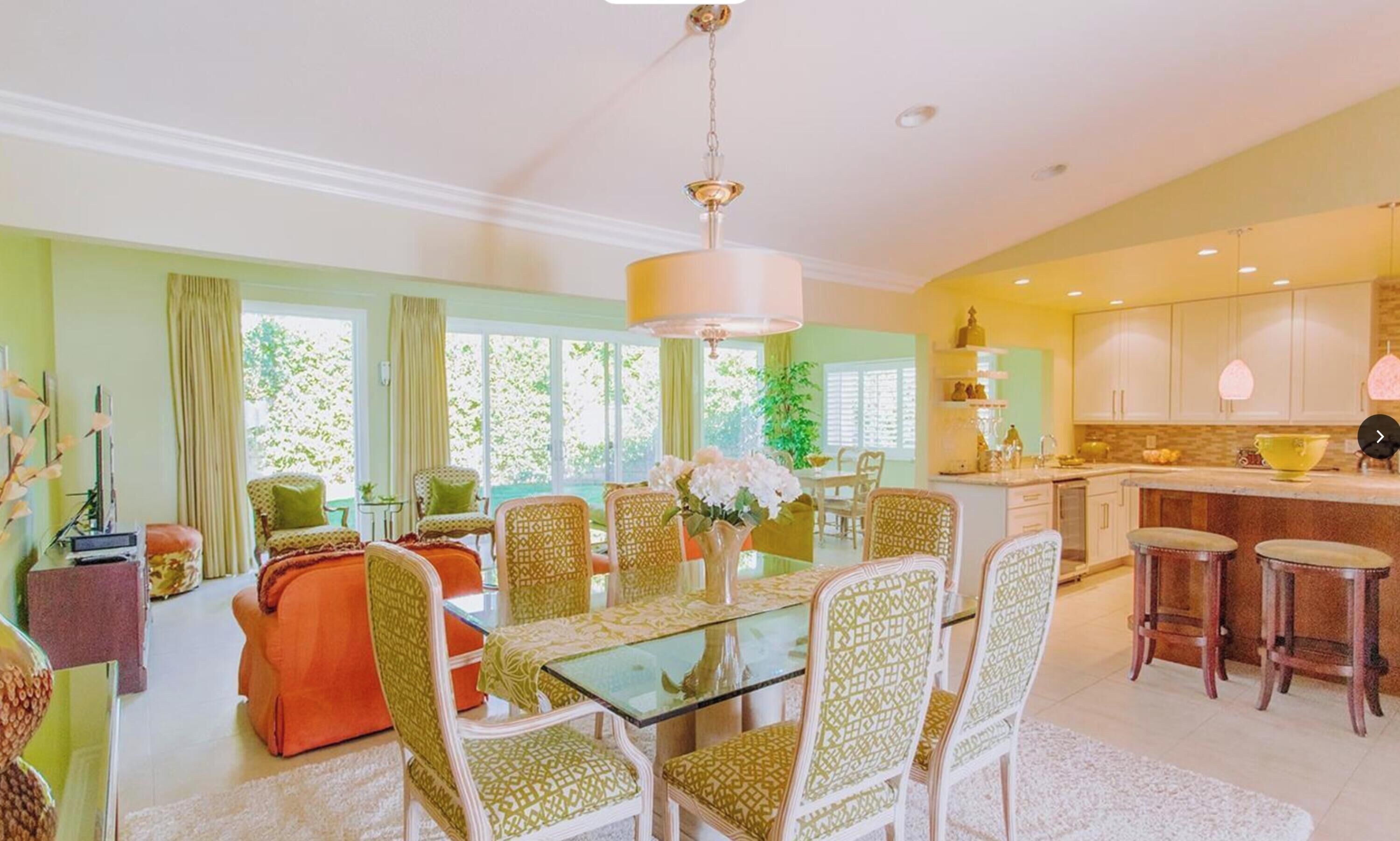 Una sala de estar amplia con una mesa de comedor de vidrio rodeada de sillas estampadas, un ventanal con cortinas y una zona de cocina con taburetes de bar.