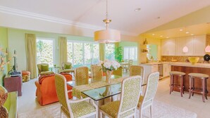 A spacious living room with a glass-top dining table surrounded by patterned chairs, a large window with curtains, and a kitchen area with bar stools. - Classic Avondale  Private  Country Club ~ Gated community front gate security (Palm Desert)