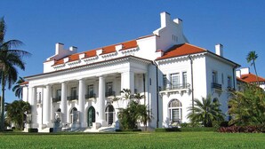 Un grand bâtiment blanc, surmonté d’un toit recouvert de tuiles rouges, avec une entrée imposante flanquée de colonnes et une pelouse bien entretenue.