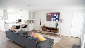 A modern living room with a grey sectional sofa, a flat-screen TV mounted on the wall, and a kitchen area in the background. - ☀️Casita bonita 🏖 Villa with 4beds/3 bath and HEATED pool 🌴🔥 (Sunrise)