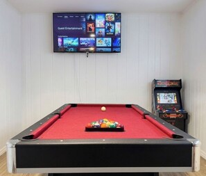 A pool table with a red felt surface, a television mounted on the wall displaying a movie selection, and a video game console to the right.