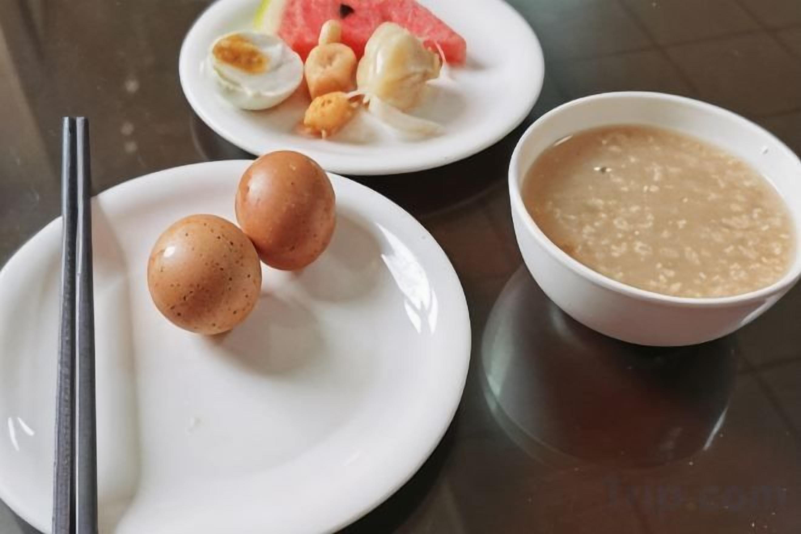 Un plato blanco con dos huevos y un par de palillos, un tazón de sopa, y un plato con una rebanada de sandía y algunas nueces.