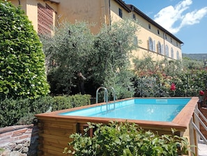Una piscina con cubierta de madera, rodeada de exuberante vegetación y un edificio con fachada amarilla.