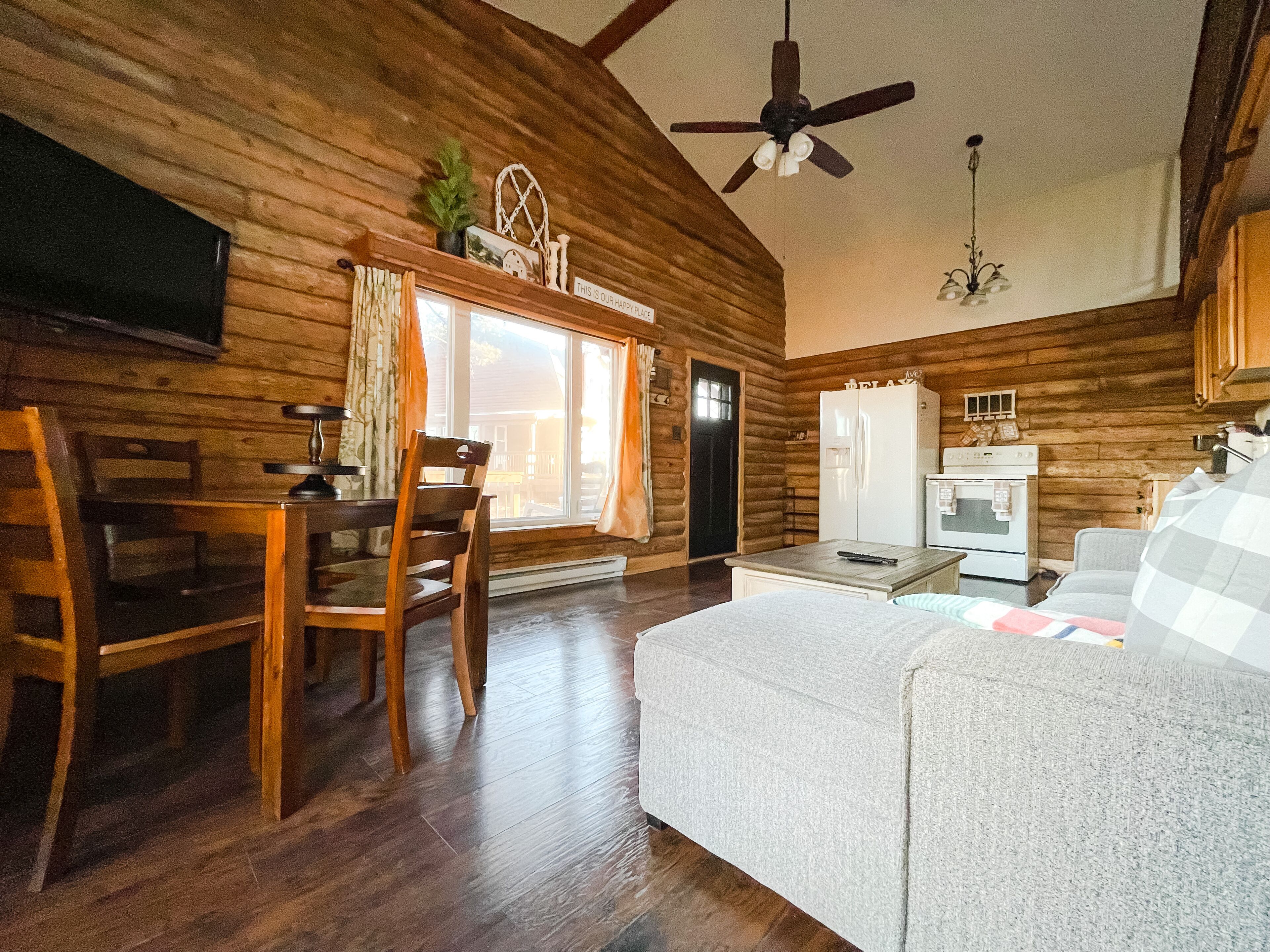 Interior acogedor de una cabaña, con paredes de madera, una zona de comedor con mesa y sillas, un sofá, un ventilador de techo y un refrigerador.