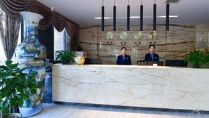 A hotel reception area with a marble counter, two receptionists, a large decorative vase, and a wall clock. - Jintai Hotel (Tongguan)