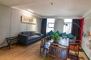 A modern living room with a grey sofa, a wooden dining table with chairs, a small side table, and a large window with curtains. - Jintai Hotel (Tongguan)