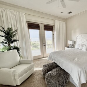 Quarto com uma cama grande, uma poltrona branca, um tapete fofinho, um ventilador de teto e vista para o oceano através de uma porta de vidro deslizante.