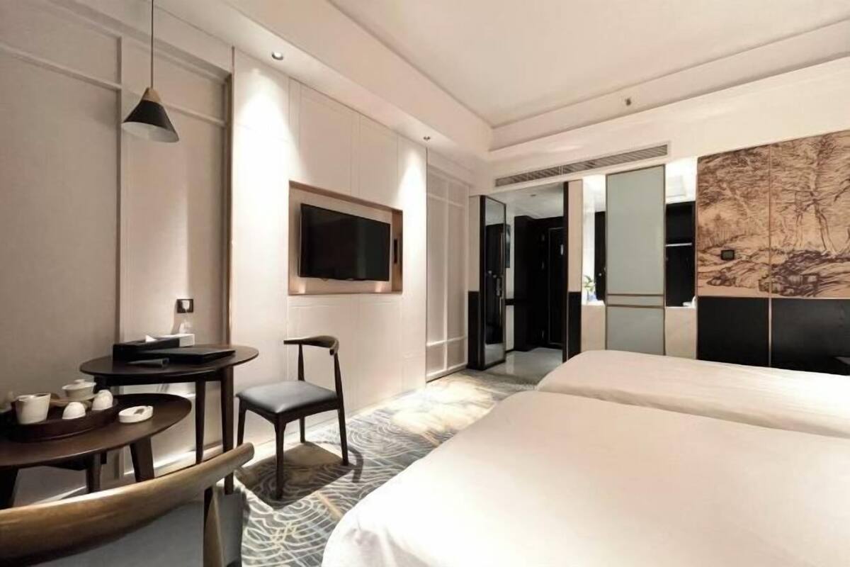 A modern hotel room with a large bed, a small round table with a chair, a TV mounted on the wall, and a large mirror.