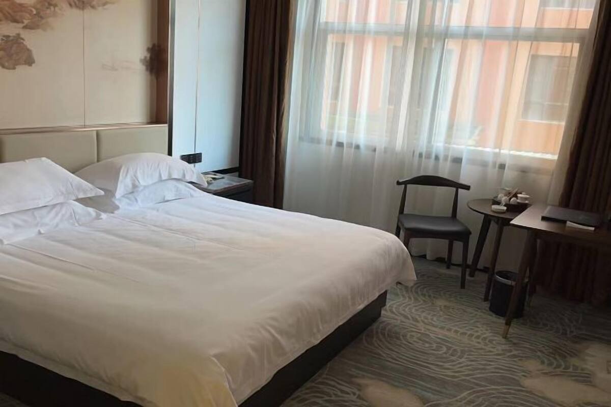 A hotel room with a large bed, a chair, a small table, and a window with sheer curtains.