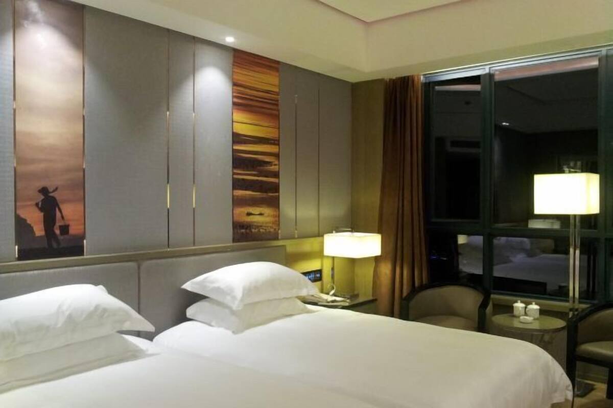 A hotel room with two beds, a nightstand with a lamp, a chair, and a large window.