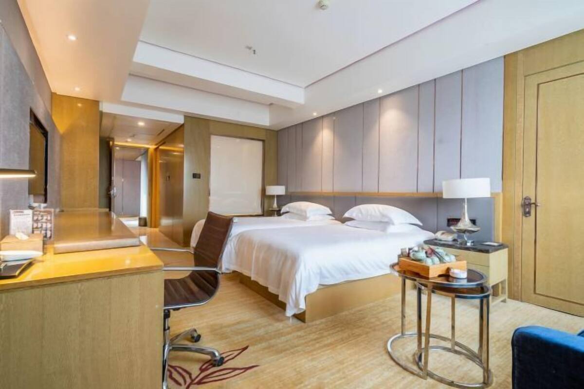 A modern hotel room with a bed, bedside table, chair, desk, and a small table with a lamp.