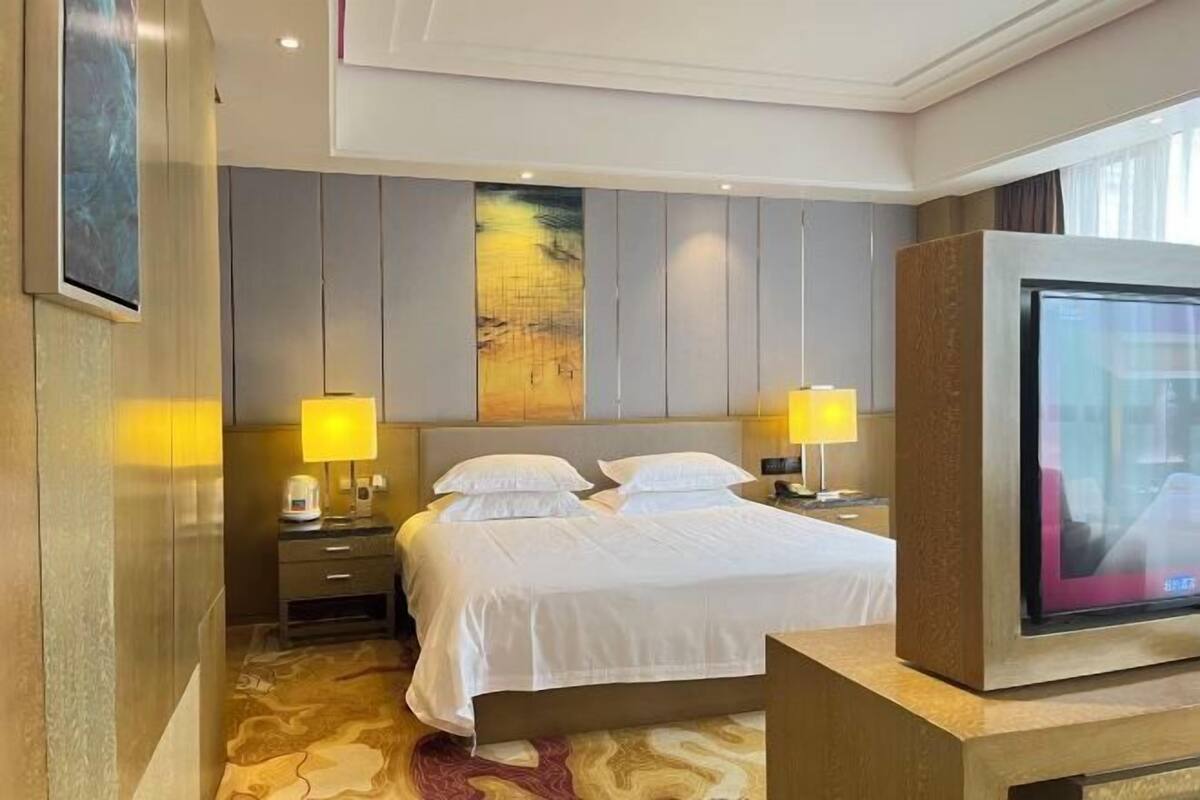 A hotel room with a bed, two bedside tables, a TV, and a large painting on the wall.