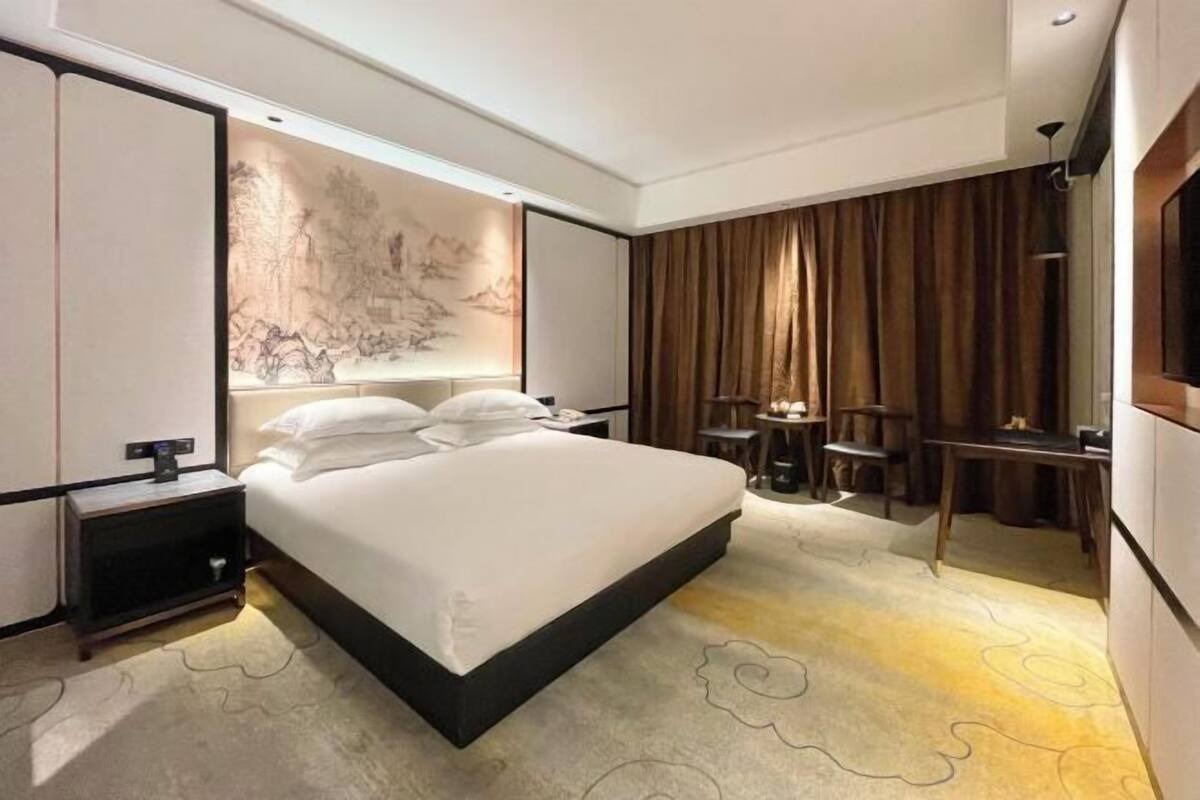 A modern hotel room with a large bed, a desk, and a chair. There is a wall-mounted TV and a small table with a lamp.