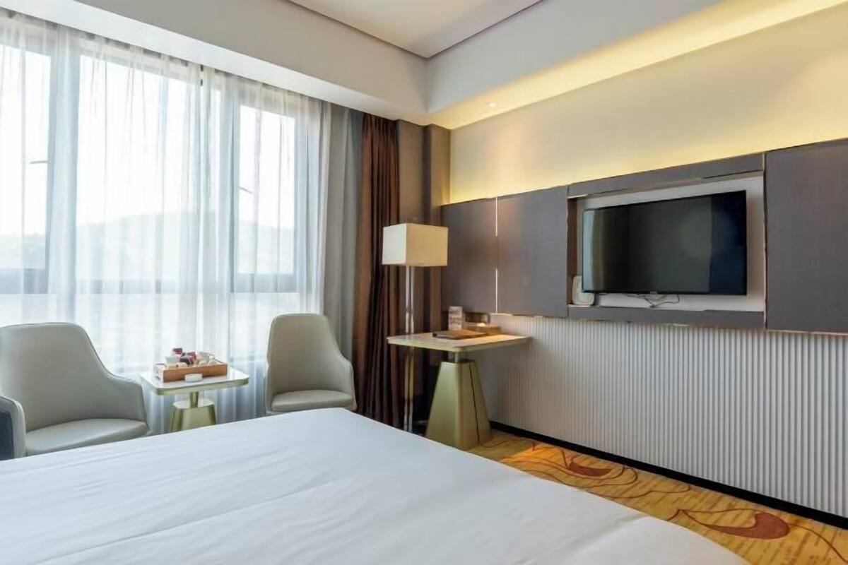 A hotel room with a large bed, a TV mounted on the wall, a small table with a lamp, two chairs, and a view of the city through large windows.