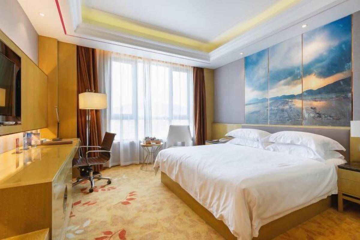 A hotel room with a bed, a desk, a chair, a TV, and a painting on the wall.
