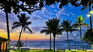 A beachside view with palm trees, a bench, and a building. - Beachfront - 2 Bedroom Suites + Air Conditioning, 2 Bathrooms, Living Room, Kitchen, 1 Garage, Wi-Fi and Bed Linen (SP)