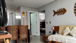 A modern living room with a sofa, a dining table with chairs, a fan, and a fish wall decoration. - Beachfront - 2 Bedroom Suites + Air Conditioning, 2 Bathrooms, Living Room, Kitchen, 1 Garage, Wi-Fi and Bed Linen (SP)