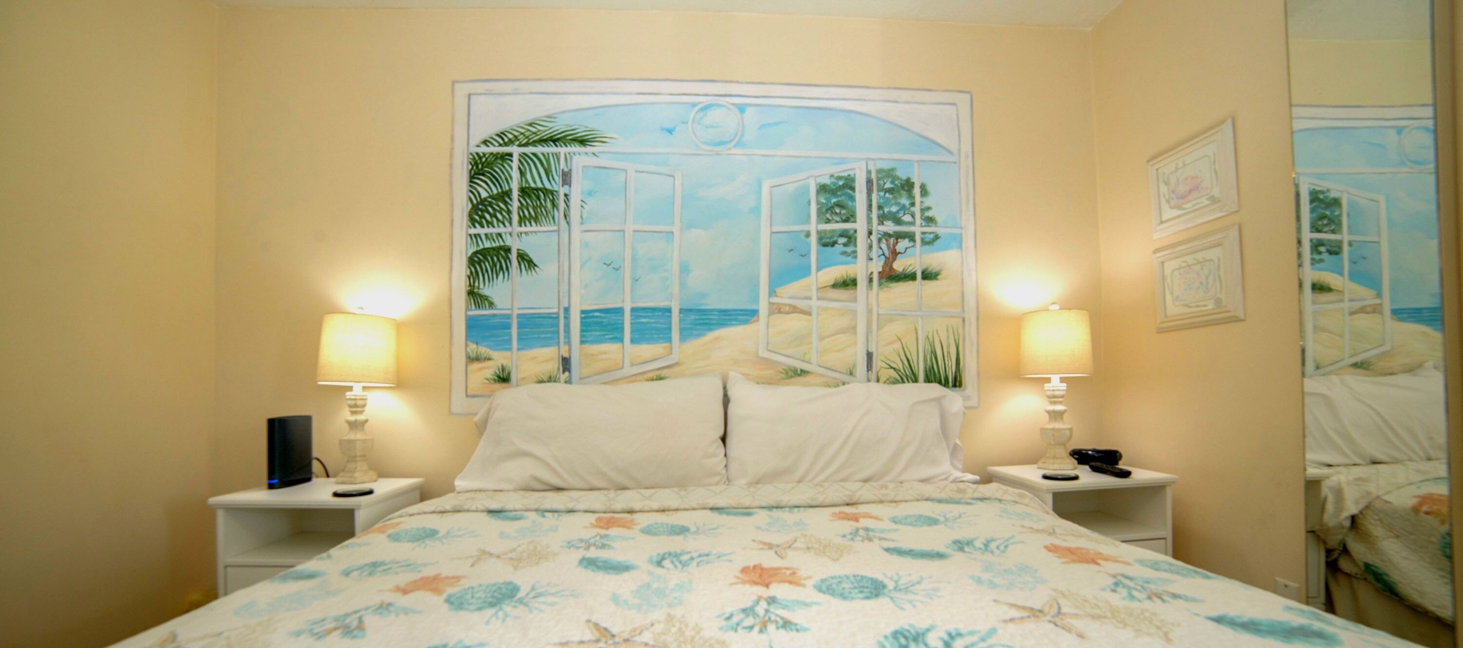 A bedroom with a bed, two bedside tables, a nightstand, a painting of a beach scene, and a mirror.