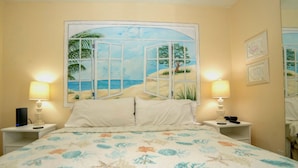 Quarto com cama, duas mesas de cabeceira, mesa de apoio, pintura de uma paisagem de praia e um espelho.
