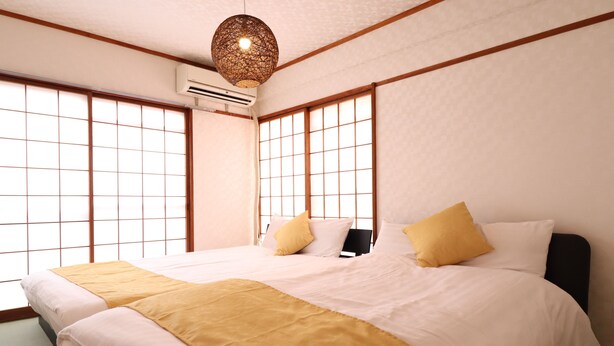 Ein traditionell japanisch gestaltetes Schlafzimmer mit Tatami-Böden, einem großen Bett mit weißer und gelber Bettwäsche sowie verschiebbaren Shoji-Schiebetüren.