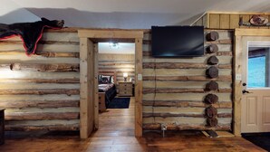 Interior de una cabaña de troncos con piso de madera, una cama con una manta roja, un televisor montado en la pared y una puerta con ventanal.