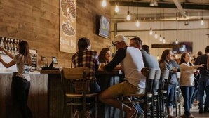 Un bar con paredes de madera y luces colgantes, gente sentada y de pie.
