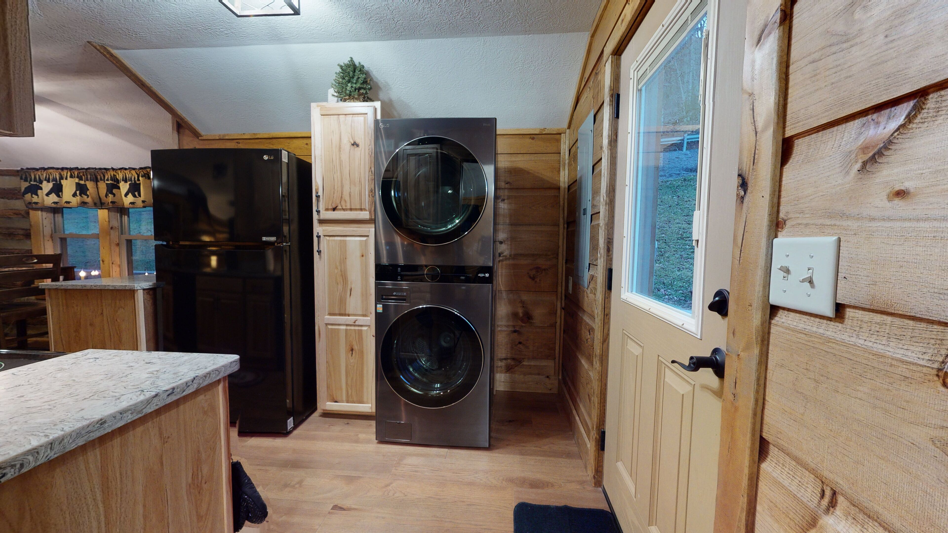 Une cuisine compacte avec un réfrigérateur noir, un appareil combiné lave-linge et sèche-linge à deux portes, des meubles de rangement en bois et une fenêtre donnant sur un espace verdoyant.