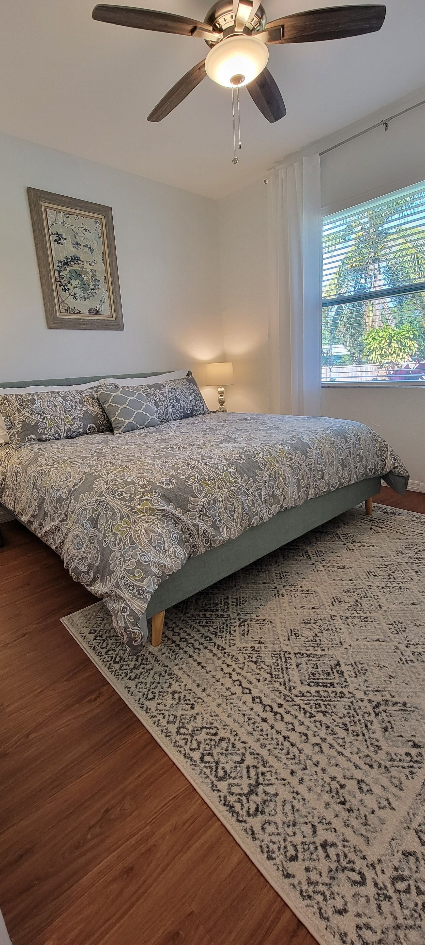 Une chambre à coucher comprenant un lit, un ventilateur de plafond, une fenêtre avec des stores et une œuvre d’art accrochée au mur.