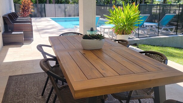 Une terrasse couverte avec une table en bois et des chaises en osier, une piscine en arrière-plan et des plantes en pot.