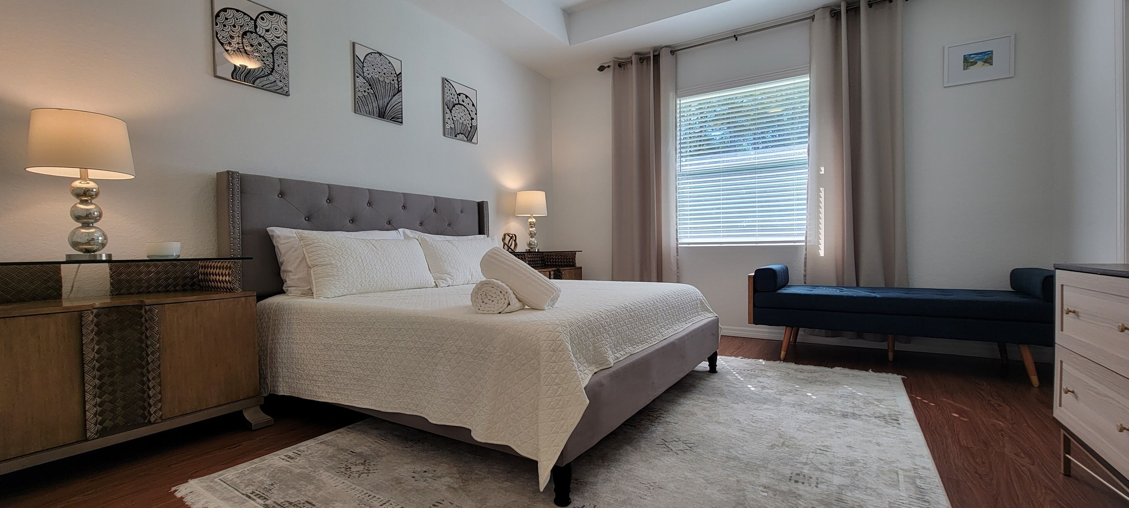 A bedroom with a large bed, bedside lamps, a bench, and artwork on the wall.
