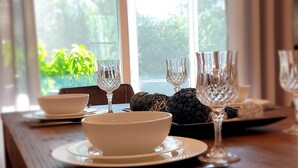Une table à manger dressée avec de l’assiette blanche, des bols et des verres en cristal, placée près d’une fenêtre ornée de rideaux.