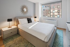 Une chambre moderne avec un grand lit, deux tables de chevet avec des lampes, un miroir fixé au mur, un radiateur et une vue sur la ville par une fenêtre.