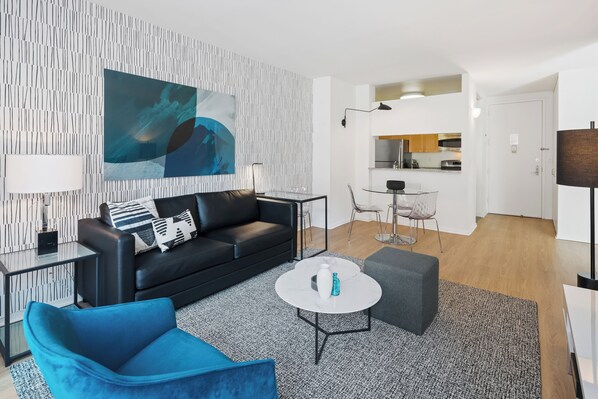 Un salon moderne avec un canapé en cuir noir, un fauteuil bleu et une table basse blanche. On aperçoit à l’arrière-plan un coin cuisine avec une table à manger et des chaises.