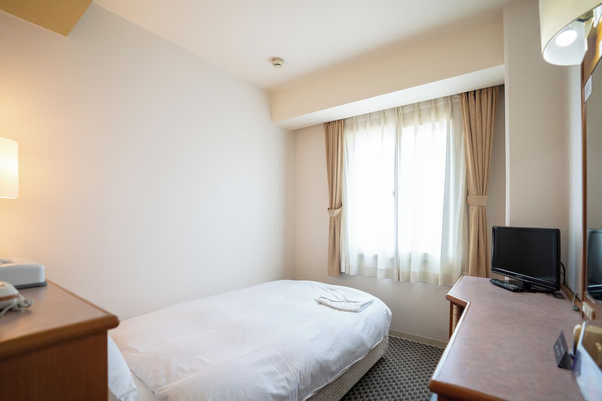 A hotel room with a single bed, a desk with a computer, a television, and a window with curtains.