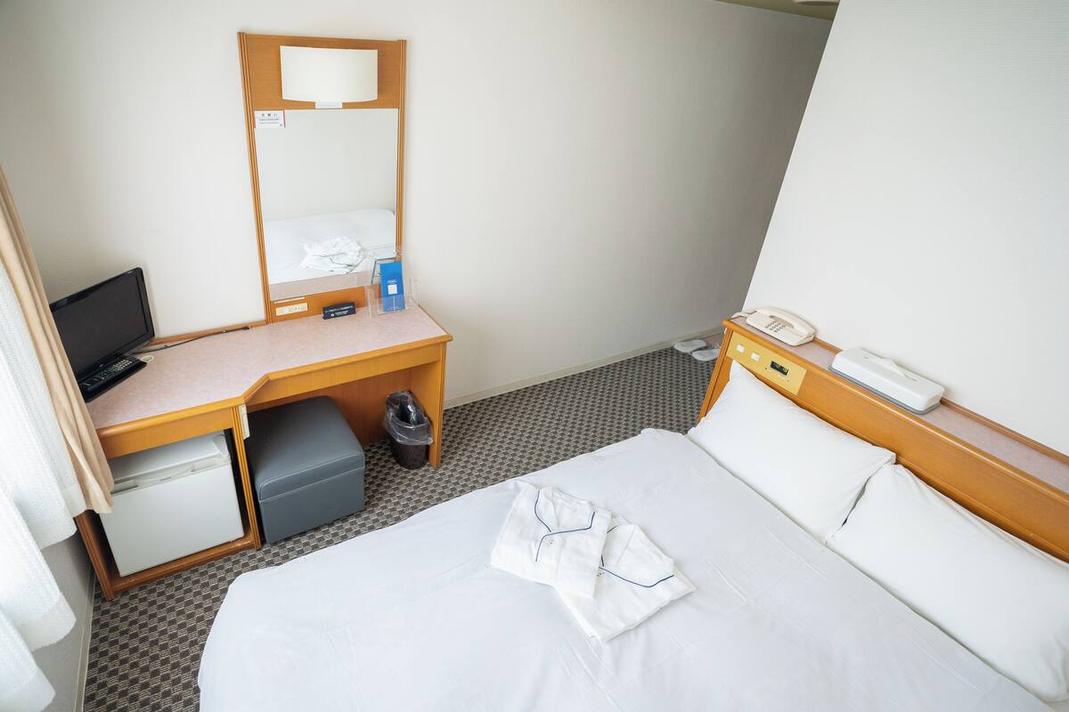 A hotel room with a bed, a desk with a computer, a phone, and a small refrigerator.