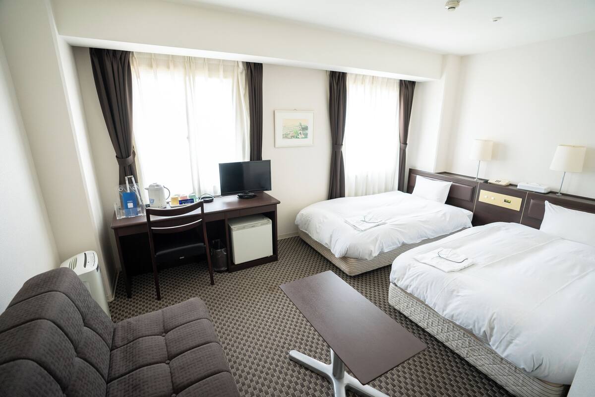A hotel room with two beds, a desk with a television, a chair, a small table, and a window with curtains.