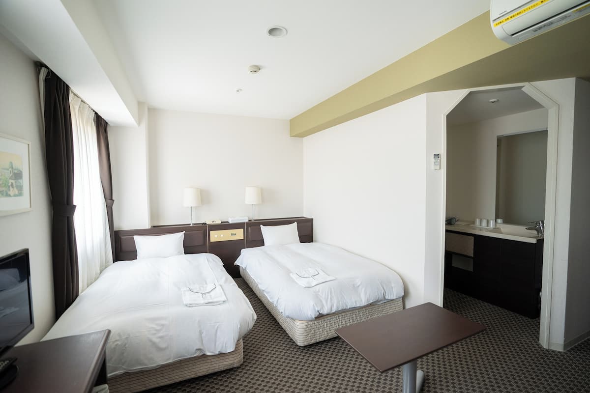 A hotel room with two beds, a desk, a chair, a TV, a bathroom with a sink and mirror, and a patterned carpet.