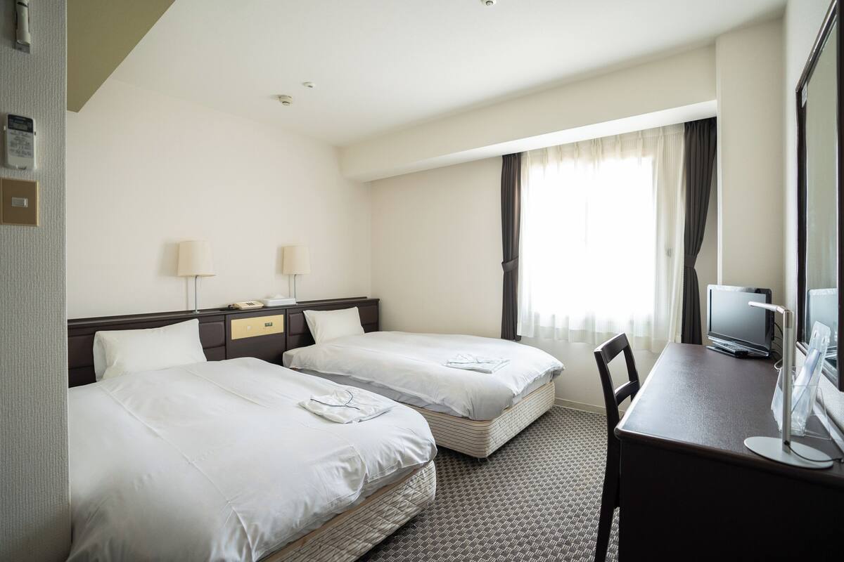 A hotel room with two single beds, a desk with a computer, and a large window with curtains.