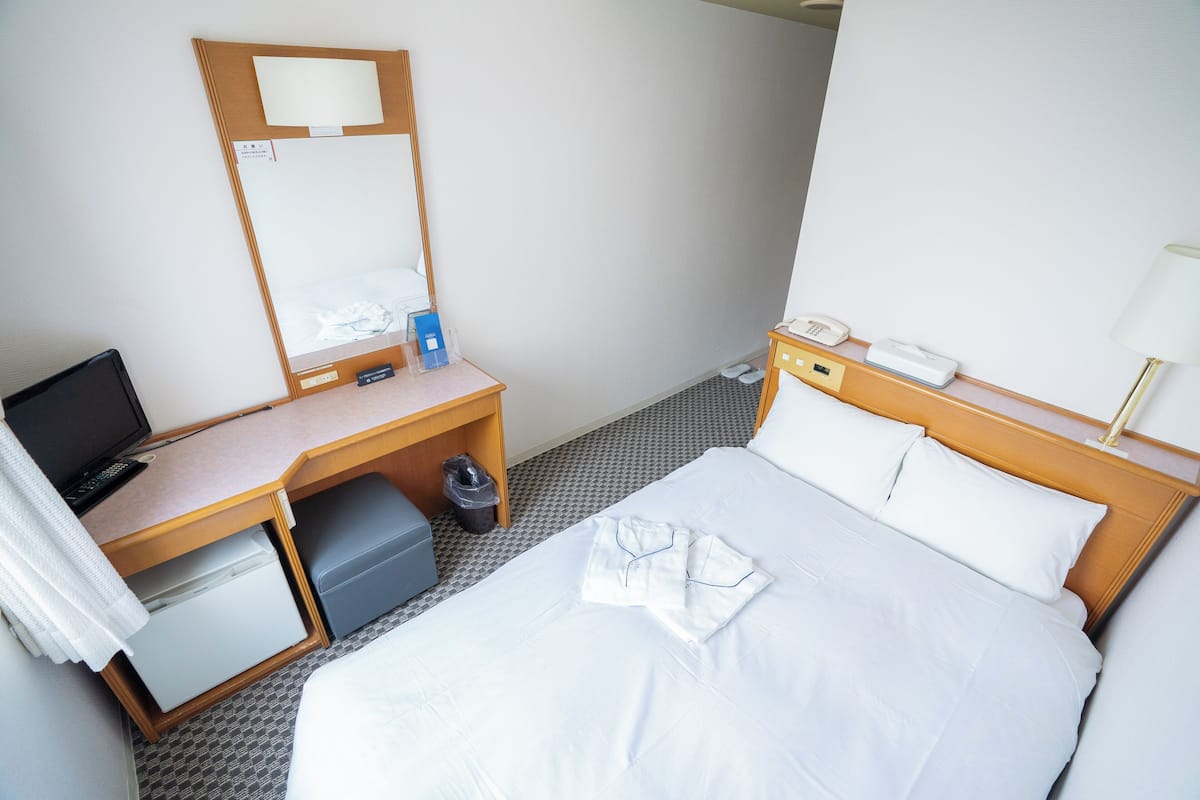 A hotel room with a bed, a desk with a computer, a small refrigerator, and a trash bin.