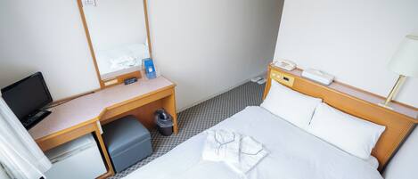 A hotel room with a bed, a desk with a computer, a small refrigerator, and a trash bin.