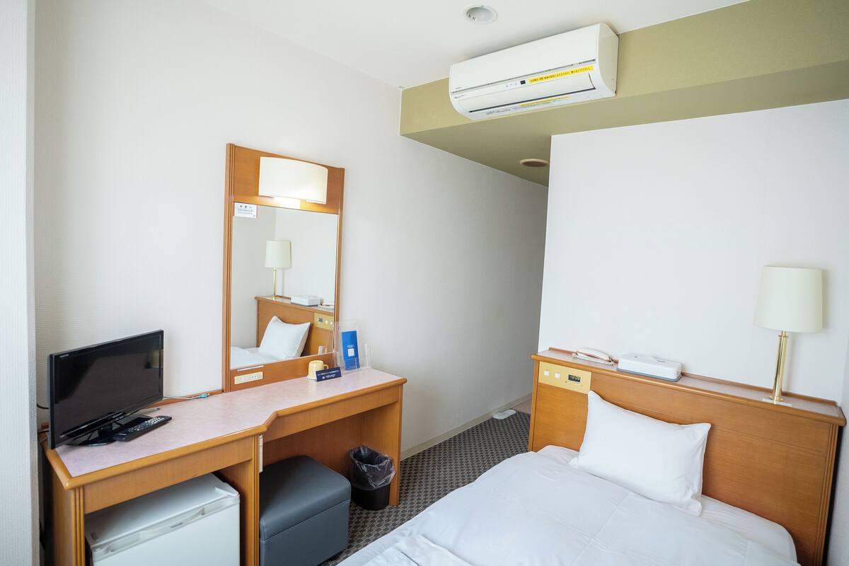 A hotel room with a bed, a desk with a computer, a chair, a trash bin, and an air conditioning unit.