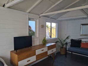 A room with a wooden dresser, a television, a window with a view, a sofa, and a potted plant. - Private cottage, dog friendly, close to beach, shops,  bush walks, fishing, more (Ettalong Beach)