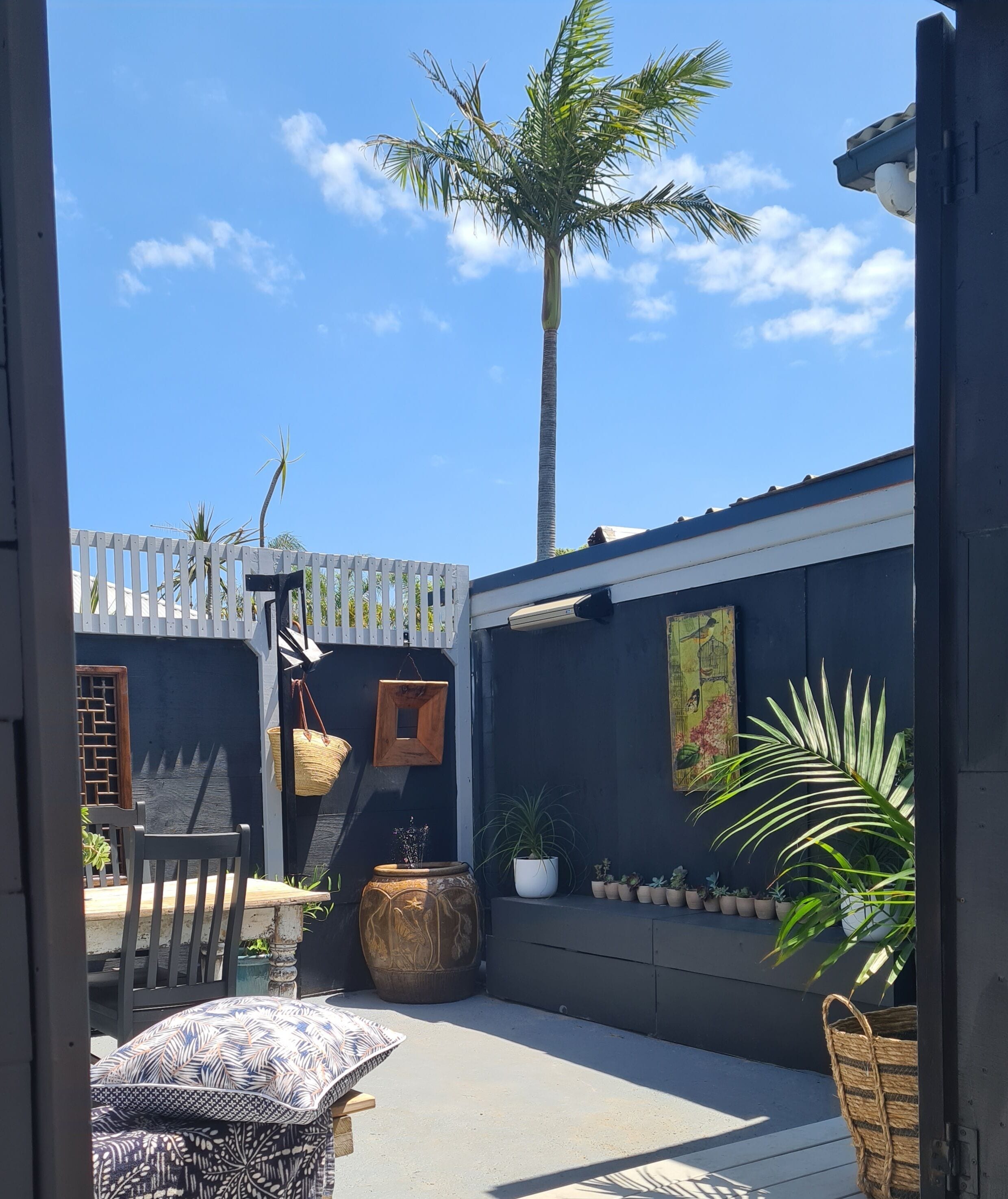 A courtyard with a wooden chair, a patterned cushion, a large decorative pot, and a variety of potted plants.