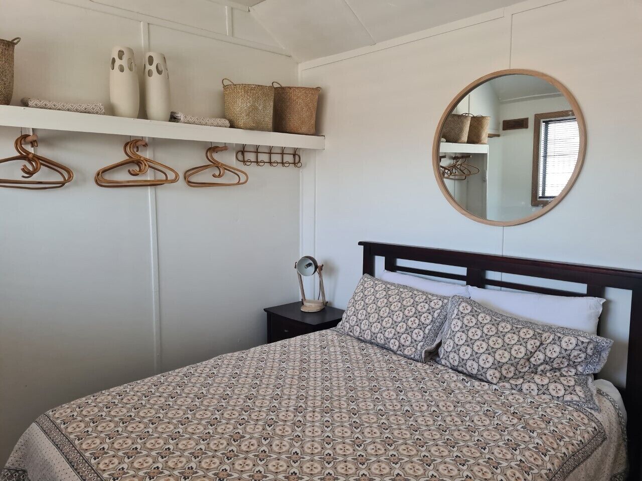 A bedroom with a patterned bedspread, a round mirror, and a bedside table with a lamp.
